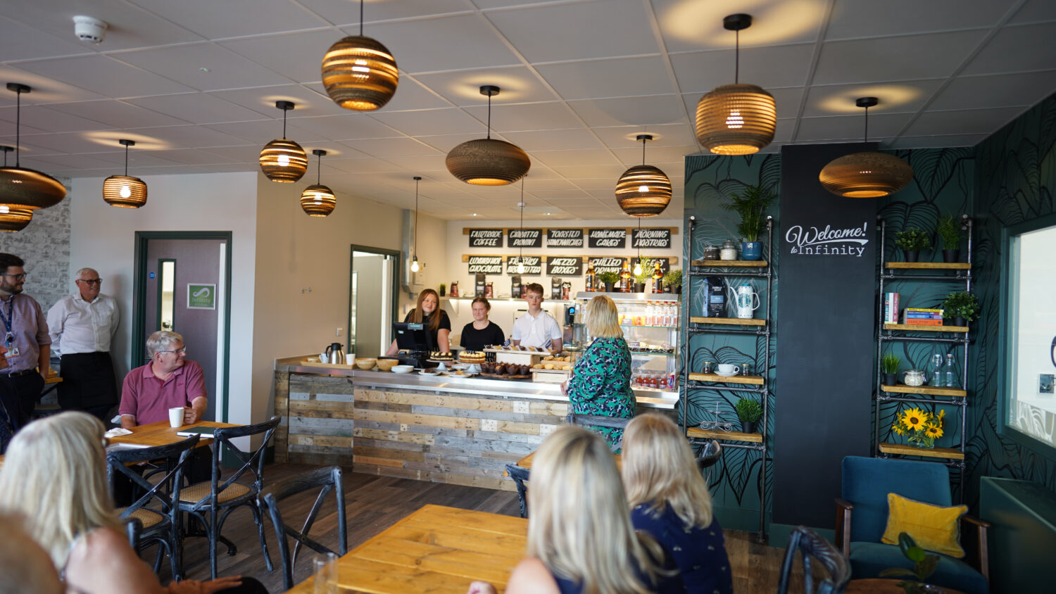 An interior photo of the Infinity Cafe at Leigh Academy Snowfields. Three staff members can be seen operating the till, with numerous guests sat at tables.