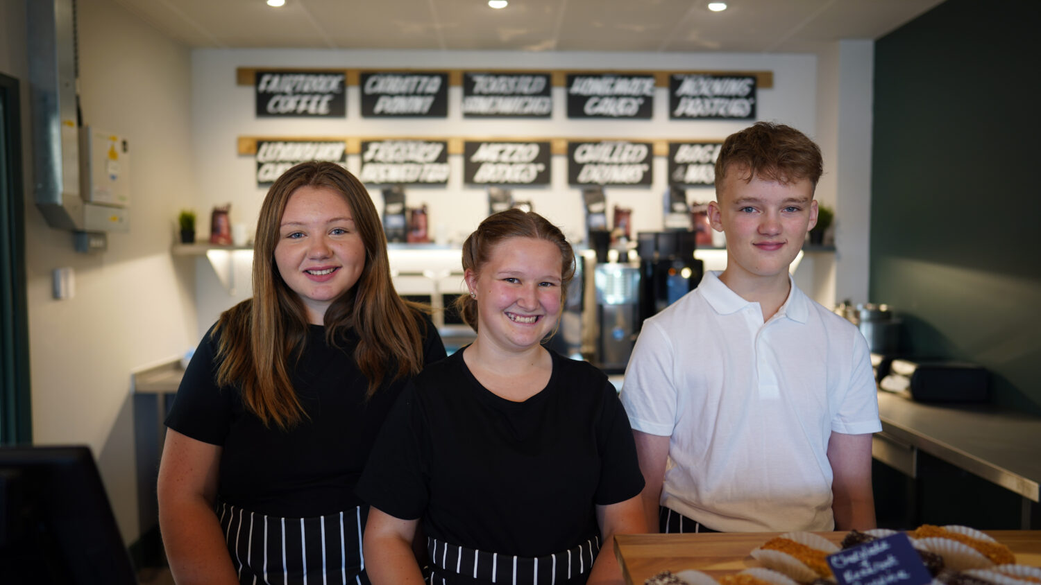 Three staff members are seen smiling together behind the till at the Infinity Cafe at Leigh Academy Snowfields.
