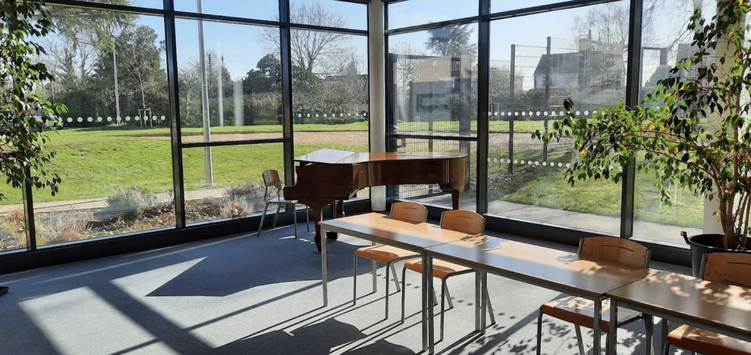 A large room with large windows, seats and a piano. The windows outlook to a green grass and trees