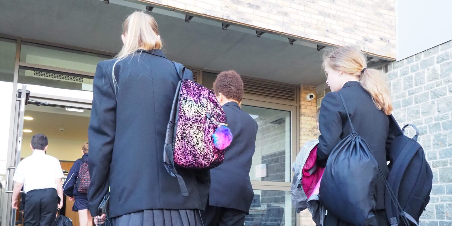 A group of students are seen entering the academy building at the start of the day with their bags on their backs.
