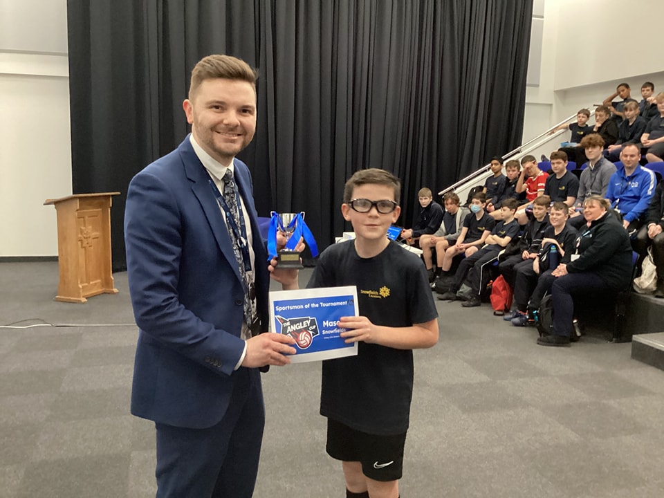 A young boy is shown being presented with a Football Tournament trophy and certificate he has won by a member of staff.