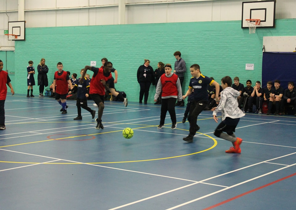 A group of boys are seen playing Football together in a Sports Hall, wearing PE Kit.