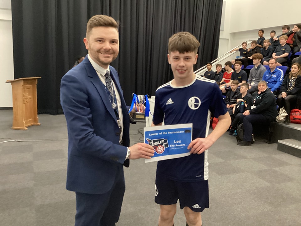 A teenage boy is shown being presented with a Football Tournament trophy and certificate he has won by a member of staff.