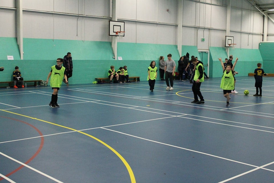 A group of Leigh Academy Snowfields students are shown wearing fluorescent bibs whilst playing a game of Football in the Sports Hall.