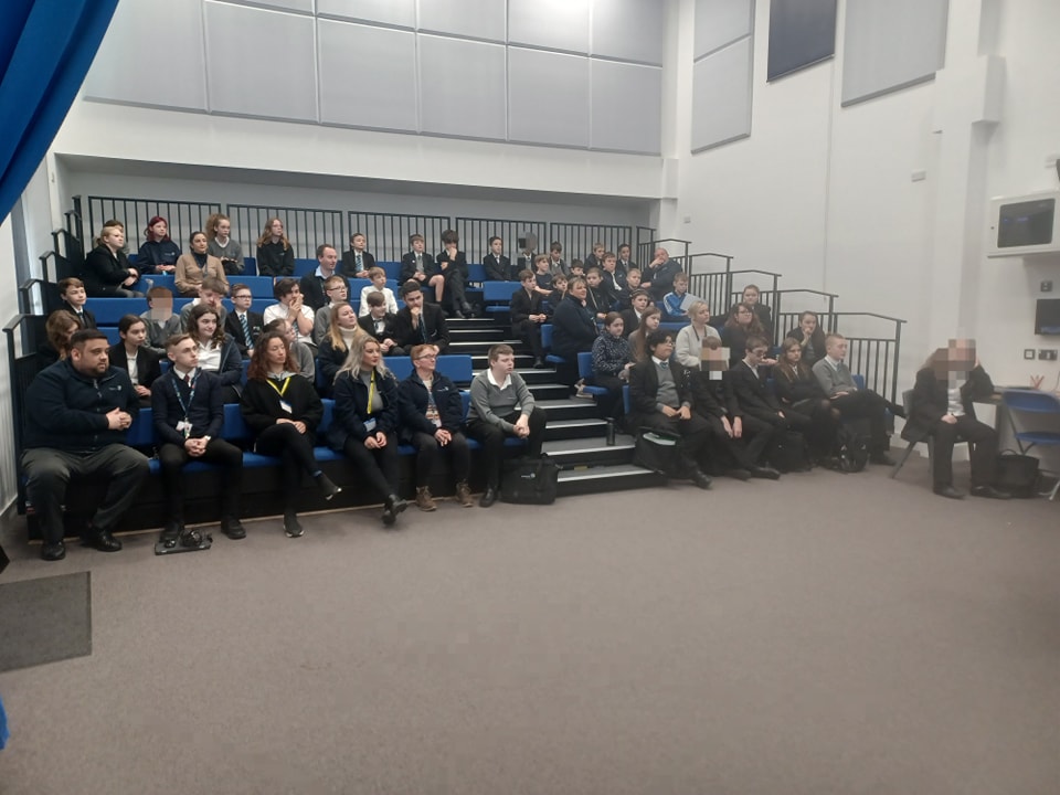 A large group of students are seen sat down together on tiered seating in the main hall for an awards presentation.