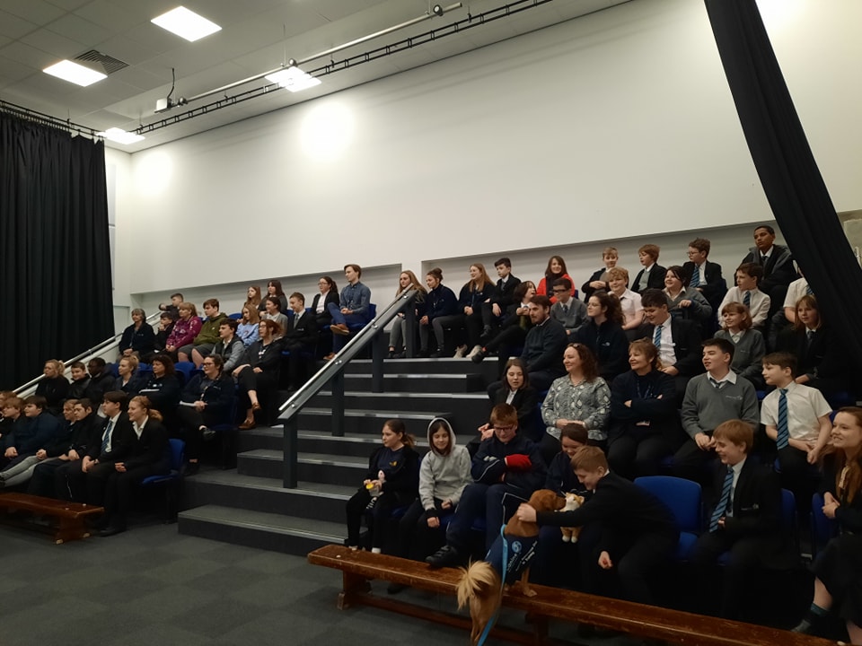 A large group of students are seen sat down together on tiered seating in the main hall for an awards presentation.