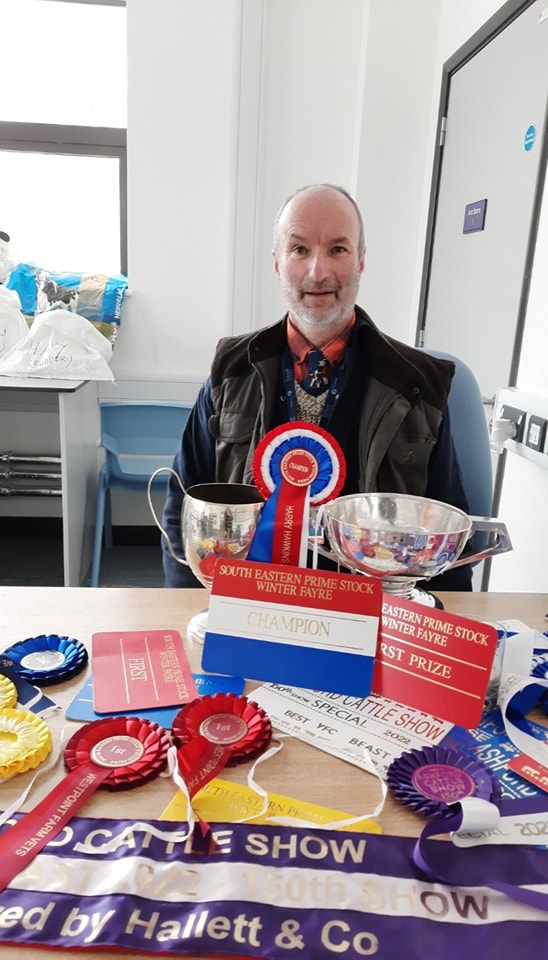 An older Caucasian man is shown sitting at a desk, smiling, with a variety of badges and trophies displayed in front of him.