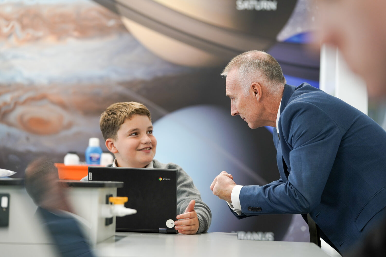 A young student can be seen working at a desk on a Chromebook laptop, whilst listening to a member of staff speaking to him on his left.