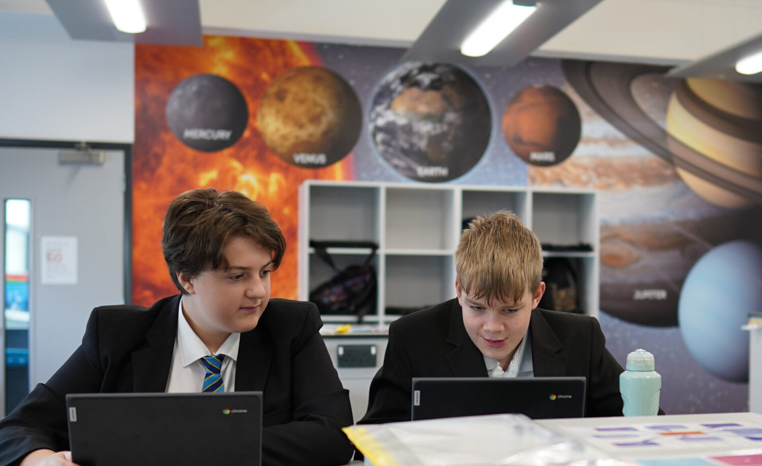 Two male students can be seen working alongside one another at a desk, using their Chromebook computers.