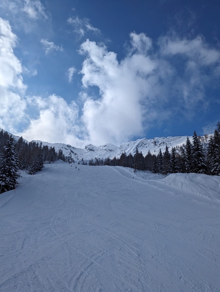 A snowy landscape pictured with mountains and trees seen in the background against a dramatic blue cloudy sky.