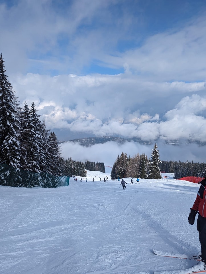 A snowy landscape is pictured with trees and mountains visible. Some Leigh Academy Snowfields students can be seen skiing down a hillside.